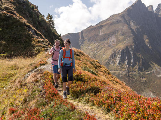Zwei Wanderer gehen auf einem schmalen Pfad in den Bergen am Spieljoch. Die Landschaft ist von buntem Herbstlaub geprägt, und im Hintergrund sind majestätische Berge zu sehen. Der Himmel ist teilweise bewölkt.