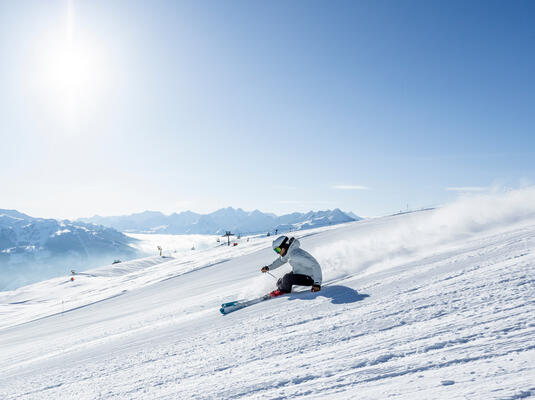 Ein Skifahrer fährt mit Schwung eine schneebedeckte Piste im Skigebiet Hochzillertal hinunter, umgeben von Bergen und einem blauen Himmel. Die Ski hinterlassen eine Schnee-Wolke und die Sonne strahlt hell.