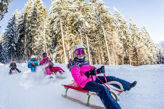 Kinder fahren auf Schlitten die Rodelbahn am Spieljoch hinunter, umgeben von Tannenbäumen. Einige tragen bunte Winterkleidung und Helme, während sie Spaß im Schnee haben. Sonnenschein reflektiert auf der glitzernden Schneedecke.