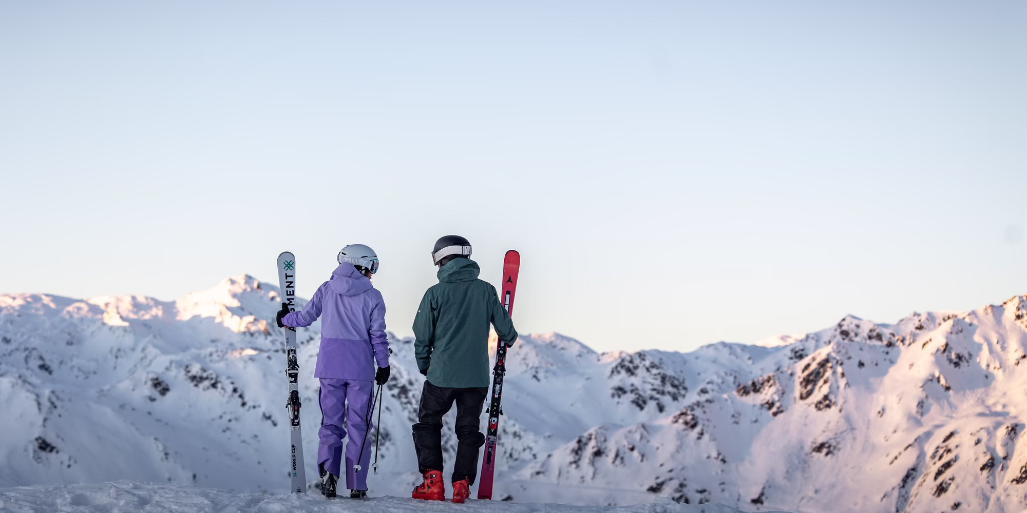 Zwei Skifahrer stehen auf einem schneebedeckten Gipfel im Zillertal und schauen auf die verschneiten Berge in der Ferne. Einer trägt eine lila Skijacke und der andere eine grüne. Beide halten ihre Skier in der Hand.