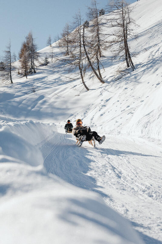 Zwei Personen fahren auf Rodeln die Rodelbahn am Spieljoch hinunter, umgeben von verschneiten Bäumen.