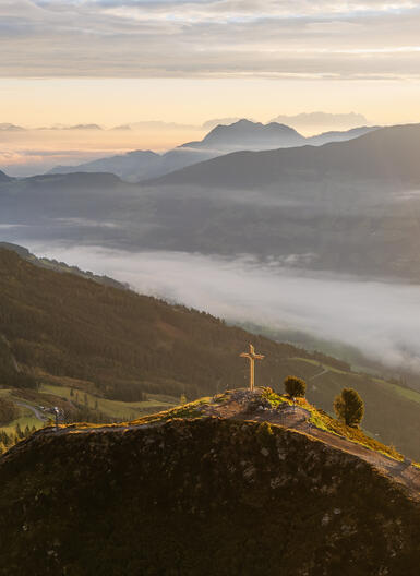 Eine malerische Aussicht auf Berge und Täler bei Sonnenaufgang. Im Vordergrund steht ein Gipfelkreuz am Spieljoch, umgeben von sanften Hügeln und Nebel, der in den Tälern schwebt. Sanfte Lichtstrahlen durchdringen die Wolken.