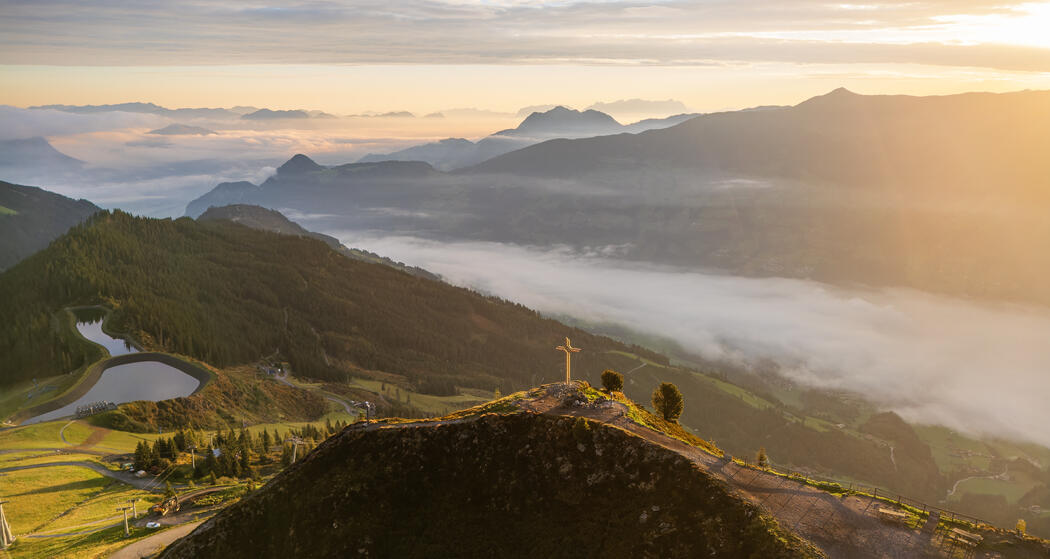Eine malerische Aussicht auf Berge und Täler bei Sonnenaufgang. Im Vordergrund steht ein Gipfelkreuz am Spieljoch, umgeben von sanften Hügeln und Nebel, der in den Tälern schwebt. Sanfte Lichtstrahlen durchdringen die Wolken.