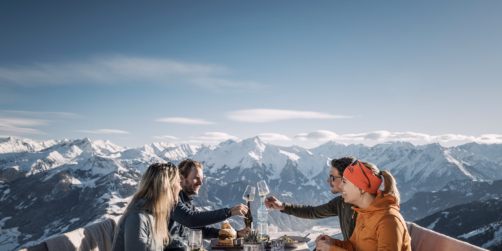 Eine Gruppe von vier Personen sitzt an einem Tisch im Freien auf der Kristallhütte. Sie stoßen mit Gläsern an und genießen ihre Speisen. Im Hintergrund sind schneebedeckte Berge und ein blauer Himmel zu sehen.