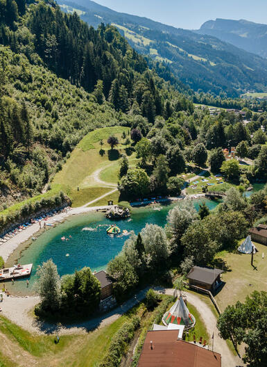 Luftaufnahme eines idyllischen Badesees in den Alpen mit umliegenden Tannenwäldern, Liegewiesen und kleinen Holzhütten.