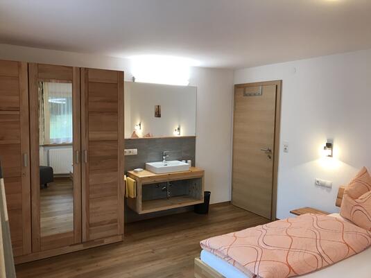 Modernes Zimmer mit Bett, Holzschrank, Waschbecken und Eingangstür. Helle Farben und freundliche Atmosphäre.
