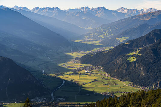 Eine weite Aussicht auf das Zillertal, umgeben von hohen Bergen. Das Tal ist grün mit landwirtschaftlichen Flächen und einem Fluss, der durch die Landschaft fließt. Die Sonne beleuchtet die Hügel und die Felsen, während die Berge im Hintergrund in Schatten liegen.