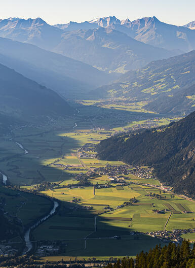 Eine weite Aussicht auf das Zillertal, umgeben von hohen Bergen. Das Tal ist grün mit landwirtschaftlichen Flächen und einem Fluss, der durch die Landschaft fließt. Die Sonne beleuchtet die Hügel und die Felsen, während die Berge im Hintergrund in Schatten liegen.