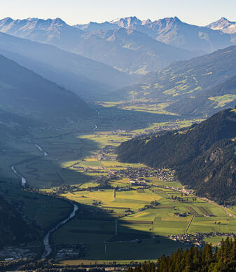 Eine weite Aussicht auf das Zillertal, umgeben von hohen Bergen. Das Tal ist grün mit landwirtschaftlichen Flächen und einem Fluss, der durch die Landschaft fließt. Die Sonne beleuchtet die Hügel und die Felsen, während die Berge im Hintergrund in Schatten liegen.