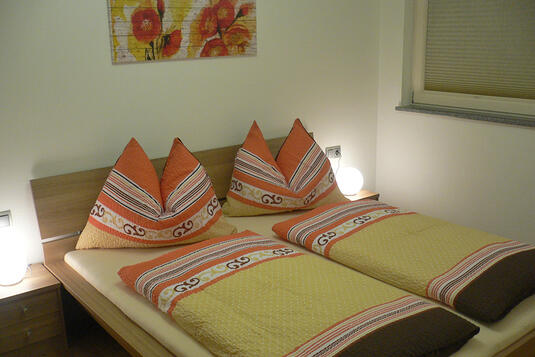 Gemütliches Schlafzimmer mit Doppelbett, dekorativen Kopfkissen und warmem Licht. Wandbild mit roten Blumen.
