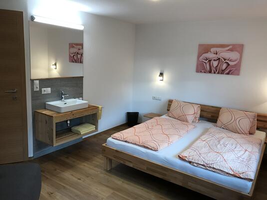 Ein modernes Schlafzimmer mit Doppelbett, Holzkommode, Waschbecken und einem dekorativen Bild an der Wand.