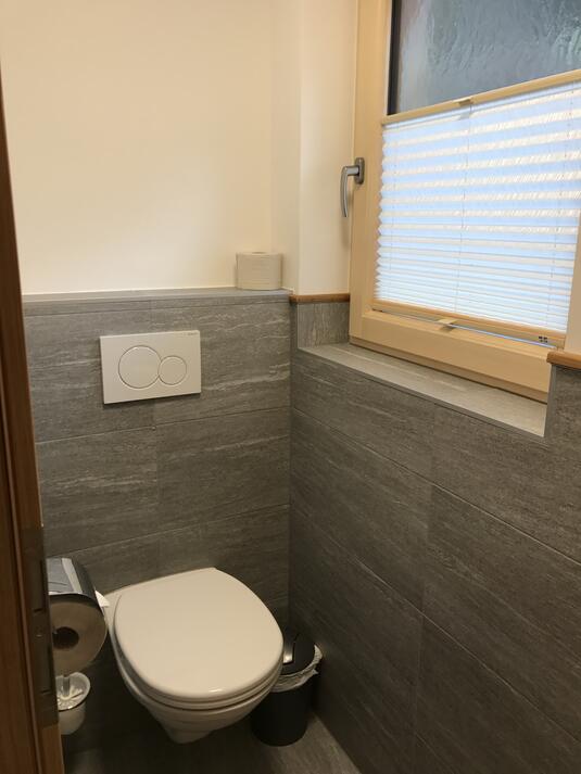 Toilette in einem kleinen Badezimmer mit grauen Fliesen und einem Fenster.