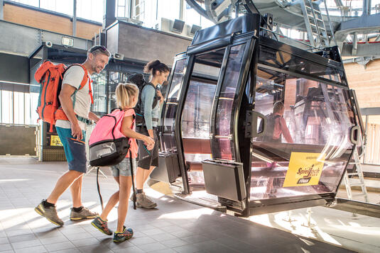 Eine Familie mit Rucksäcken steigt in eine Gondel der Spieljochbahn ein. Die Umgebung ist hell erleuchtet durch die Sonnenstrahlen. und die Familie ist fröhlich.