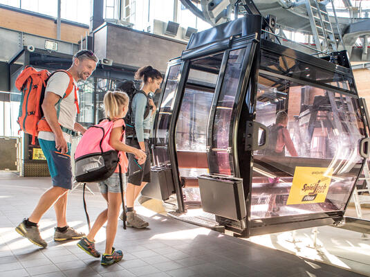 Eine Familie mit Rucksäcken steigt in eine Gondel der Spieljochbahn ein. Die Umgebung ist hell erleuchtet durch die Sonnenstrahlen. und die Familie ist fröhlich.
