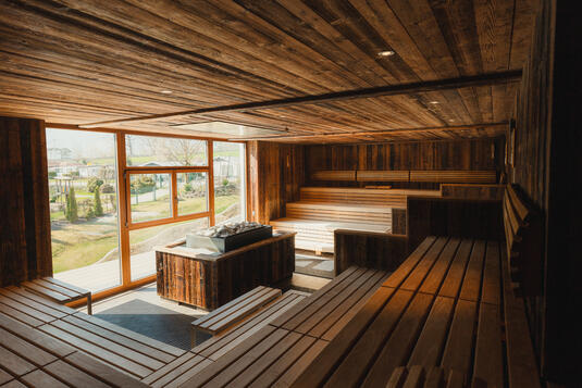 Innenansicht einer Sauna mit Holzwänden und -bänken. Große Fenster lassen Tageslicht herein und bieten einen Blick ins Freie. Die Umgebung ist grün und einladend.