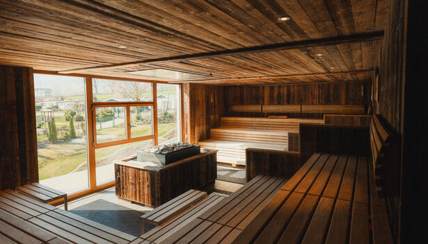 Innenansicht einer Sauna mit Holzwänden und -bänken. Große Fenster lassen Tageslicht herein und bieten einen Blick ins Freie. Die Umgebung ist grün und einladend.