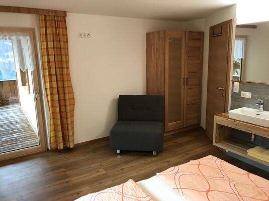 Modernes Zimmer mit Holzboden, grauem Sessel, Bett und Fenster mit Blick auf eine Terrasse.