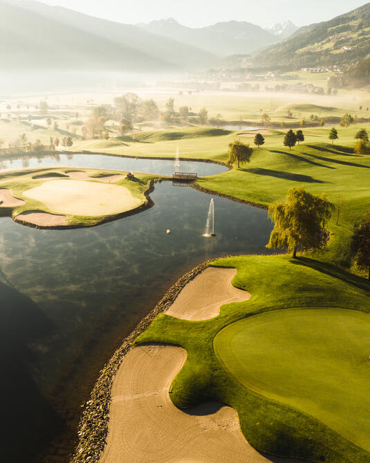 Eine Aussicht auf den Golfplatz in Uderns im Zillertal mit gepflegten Fairways und Sandbunkern, die von Wasserflächen umgeben sind. Die Landschaft wird von sanften Hügeln und Bergen im Hintergrund geschmückt. Es ist Nebel am Morgen zu sehen, der eine ruhige Atmosphäre schafft.