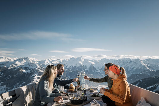 Eine Gruppe von vier Personen sitzt an einem Tisch im Freien auf der Kristallhütte. Sie stoßen mit Gläsern an und genießen ihre Speisen. Im Hintergrund sind schneebedeckte Berge und ein blauer Himmel zu sehen.