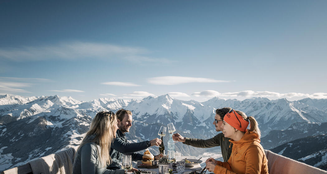 Eine Gruppe von vier Personen sitzt an einem Tisch im Freien auf der Kristallhütte. Sie stoßen mit Gläsern an und genießen ihre Speisen. Im Hintergrund sind schneebedeckte Berge und ein blauer Himmel zu sehen.
