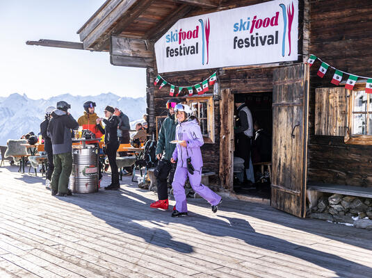 Skifood Festival mit Gästen vor einer Berghütte, umgeben von Bergen und festlicher Dekoration.