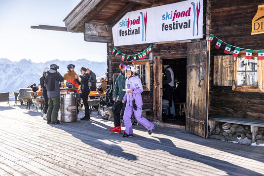 Skifood Festival mit Gästen vor einer Berghütte, umgeben von Bergen und festlicher Dekoration.