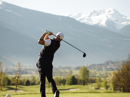 Ein Golfer schlägt einen Ball auf dem Platz im Zillertal mit Bergen im Hintergrund und klarem Himmel.