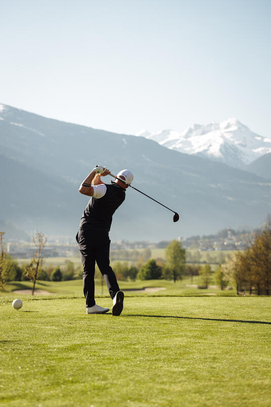 Ein Golfer schlägt einen Ball auf dem Platz im Zillertal mit Bergen im Hintergrund und klarem Himmel.