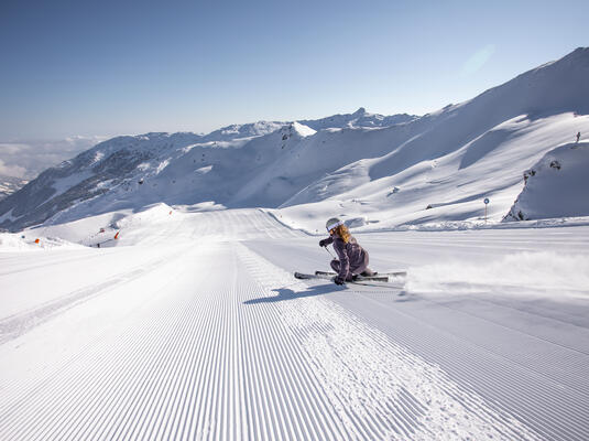 Eine Skifahrerin gleitet über eine frisch präparierte Piste in den Bergen im Skigebiet Hochfügen. Die Umgebung ist von schneebedeckten Gipfeln umgeben und der Himmel ist klar und sonnig. Die Piste zieht sich in die Ferne.