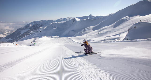 Eine Skifahrerin gleitet über eine frisch präparierte Piste in den Bergen im Skigebiet Hochfügen. Die Umgebung ist von schneebedeckten Gipfeln umgeben und der Himmel ist klar und sonnig. Die Piste zieht sich in die Ferne.