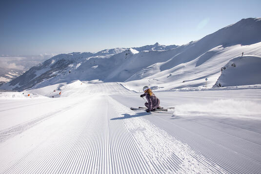 Eine Skifahrerin gleitet über eine frisch präparierte Piste in den Bergen im Skigebiet Hochfügen. Die Umgebung ist von schneebedeckten Gipfeln umgeben und der Himmel ist klar und sonnig. Die Piste zieht sich in die Ferne.