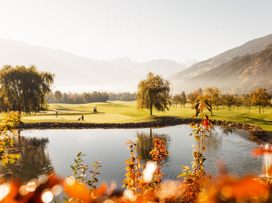 Eine malerische Golfplatzlandschaft am Golfclub Zillertal mit sanften Hügeln, Bäumen und einem Teich im Vordergrund. Im Hintergrund spielen Golfer. Die Szenerie wird von herbstlichen Farben und klarem Himmel begleitet.