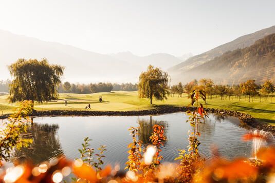 Eine malerische Golfplatzlandschaft am Golfclub Zillertal mit sanften Hügeln, Bäumen und einem Teich im Vordergrund. Im Hintergrund spielen Golfer. Die Szenerie wird von herbstlichen Farben und klarem Himmel begleitet.
