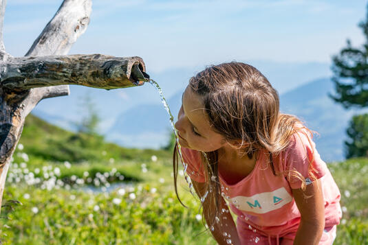 Ein Mädchen beugt sich über einen Wasserauslauf in einer grünen Berglandschaft und trinkt frisches Wasser.