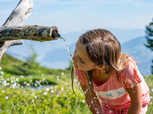 Ein Mädchen beugt sich über einen Wasserauslauf in einer grünen Berglandschaft und trinkt frisches Wasser.
