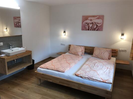 Modernes Schlafzimmer mit Holzbetten, zwei Kissen, Waschbecken und Wandspiegel im Hintergrund.